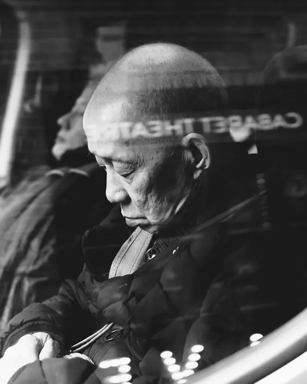 An older man rests his head by a vehicle window, in black and white.