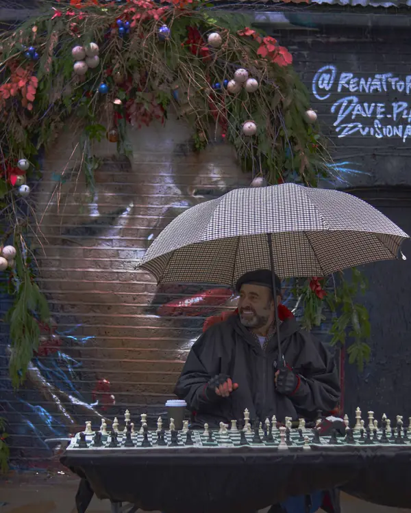 A man sits beneath an umbrella beside a street chess set and a painted mural.