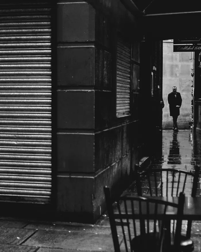 A lone figure walks past a closed shutter on a narrow London street, in black and white.